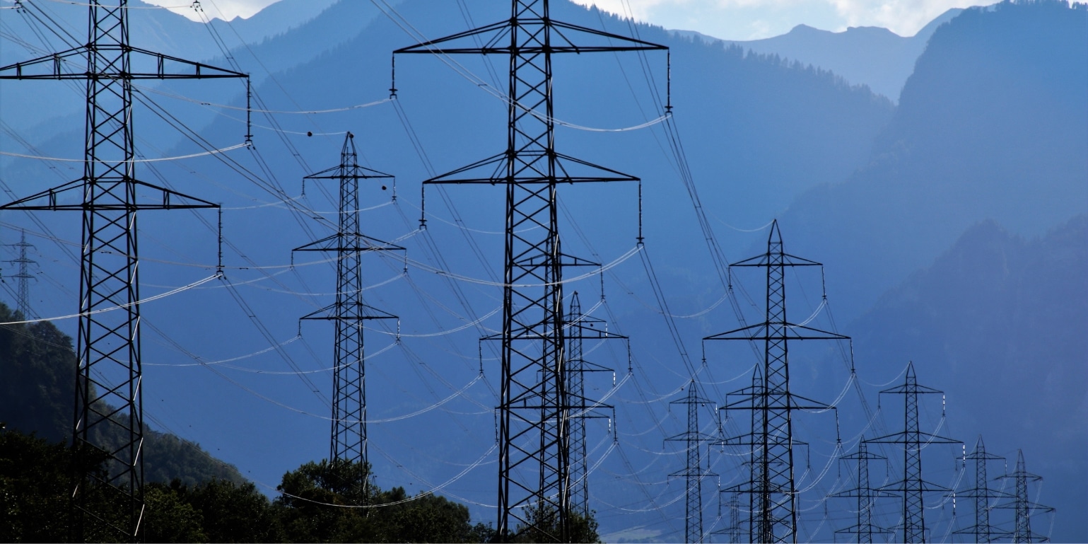 Blue hued landscape photo of a series of power lines into the distance, with tree covered mountains in the background. Used to represent the concept of procurement technology connecting organisations to a network of suppliers.