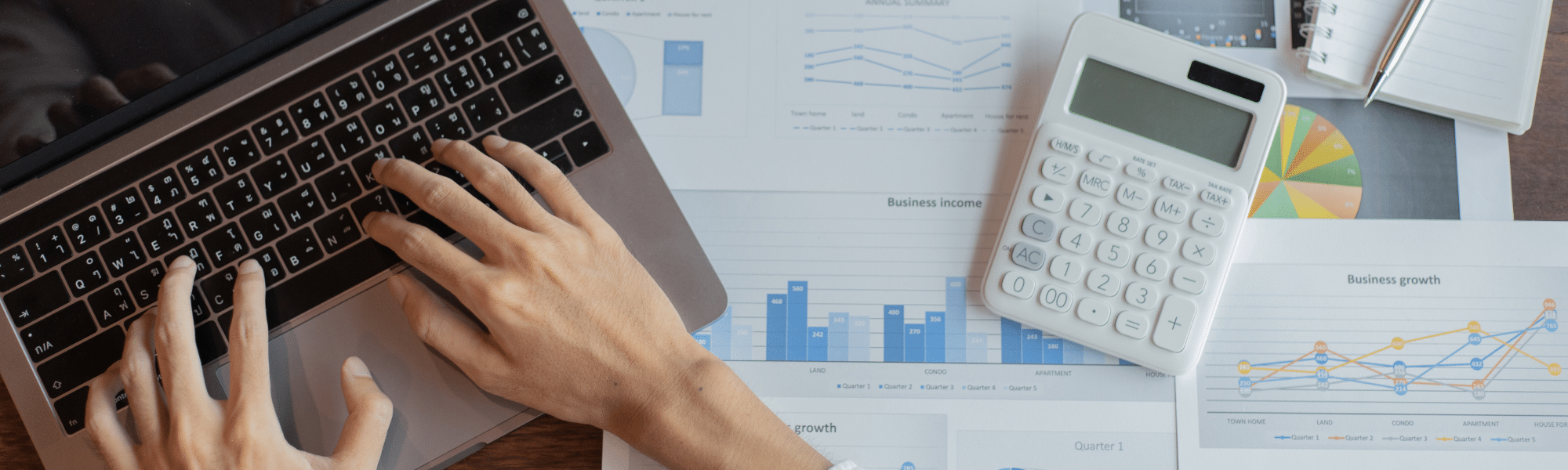 A blog feature image of a hand typing on a laptop on a desk. The desk is covered with sheets of paper containing business data and a calculator. This represents staff from the finance department, specifically in the AP or AR function struggling with manual invoice processing and in dire need of invoice processing automation.