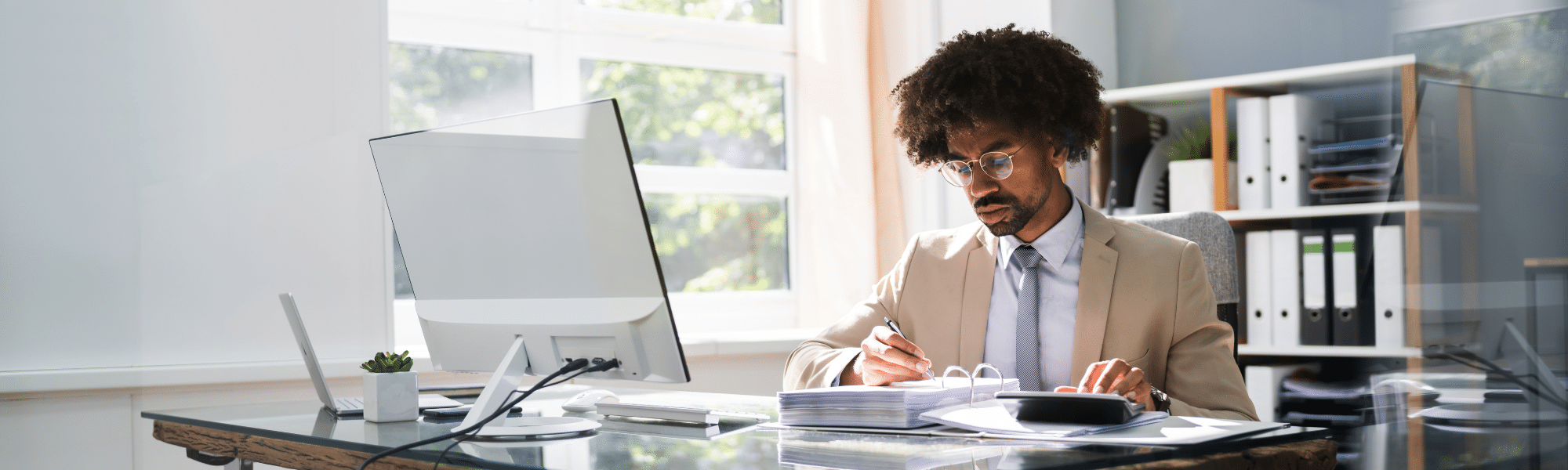 A blog feature image of a young black male finance executive sat at his office desk reviewing accounts processing manually. He represents a finance staff, whether that be in the AP and/or AR function, considering invoice processing automation.