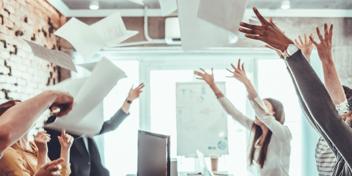 A blog feature image depicts a group of workers in an office, jubilantly throwing sheets of paper in the air. Their celebration signifies the shift from traditional invoicing to the use of eInvoicing through Accounts Receivable automation software.