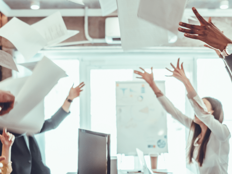 A blog feature image depicts a group of workers in an office, jubilantly throwing sheets of paper in the air. Their celebration signifies the shift from traditional invoicing to the use of eInvoicing through Accounts Receivable automation software.