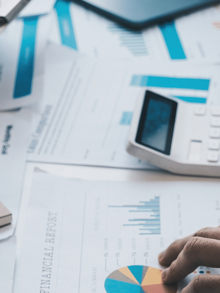 Blog feature image showing a close up of a messy desk with scattered papers; one pair of hands holds a clipboard and another pair of hands use a calculator. It represents the AP and AR teams working hard to fulfil the invoicing process.