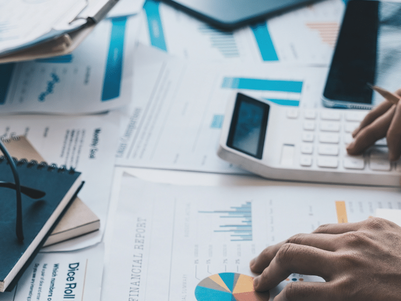 Blog feature image showing a close up of a messy desk with scattered papers; one pair of hands holds a clipboard and another pair of hands use a calculator. It represents the AP and AR teams working hard to fulfil the invoicing process.