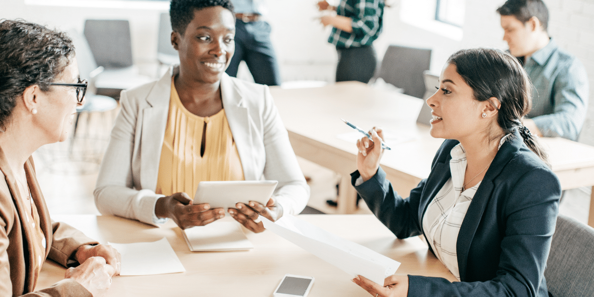 A blog feature image shows three female accountants in an office, discussing work whilst holding electronic gadgets. They sit around a desk filled with invoices, symbolising the shift from paper invoices to electronic invoicing software in the AP and AR team.