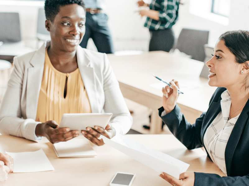 A blog feature image shows three female accountants in an office, discussing work whilst holding electronic gadgets. They sit around a desk filled with invoices, symbolising the shift from paper invoices to electronic invoicing software in the AP and AR team.