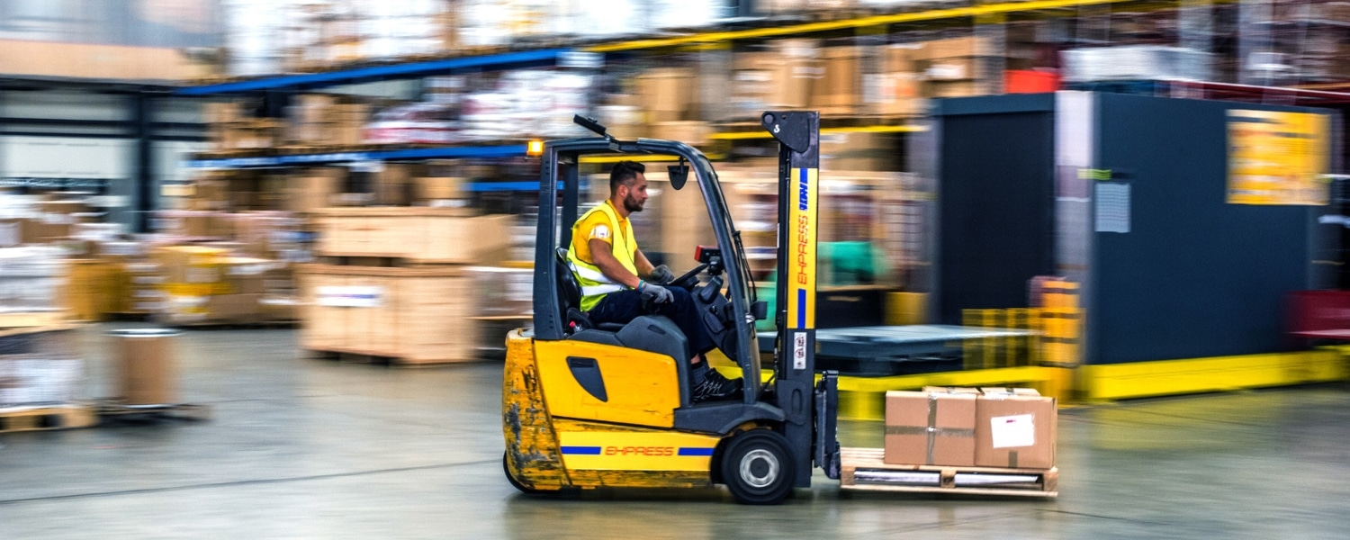 Warehouse worker in high visibility jacket operating a forklift. Surroundings of the warehouse are blurred, implying the forklift is in fast forward-motion. Representing blog titled: The real EDI challenges businesses face with traditional systems