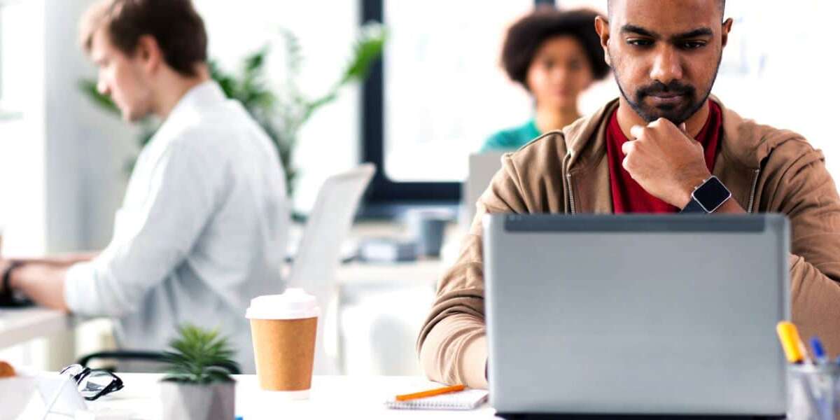 Male executive sits in modern office environment with laptop and takeaway coffee studying his screen looking perplexed. Two colleagues sit on separate desks behind him working on their laptops. Representing blog titled: Why your business needs a modern EDI platform