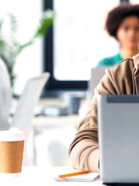 Male executive sits in modern office environment with laptop and takeaway coffee studying his screen looking perplexed. Two colleagues sit on separate desks behind him working on their laptops. Representing blog titled: Why your business needs a modern EDI platform