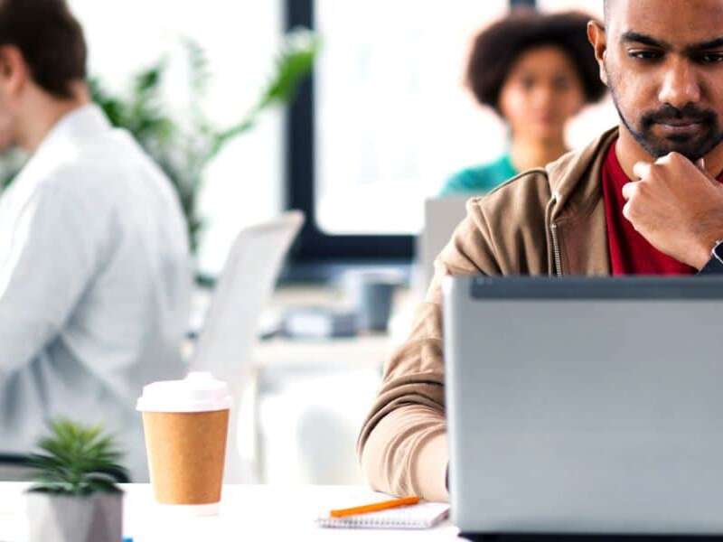 Male executive sits in modern office environment with laptop and takeaway coffee studying his screen looking perplexed. Two colleagues sit on separate desks behind him working on their laptops. Representing blog titled: Why your business needs a modern EDI platform