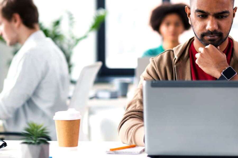 Male executive sits in modern office environment with laptop and takeaway coffee studying his screen looking perplexed. Two colleagues sit on separate desks behind him working on their laptops. Representing blog titled: Why your business needs a modern EDI platform