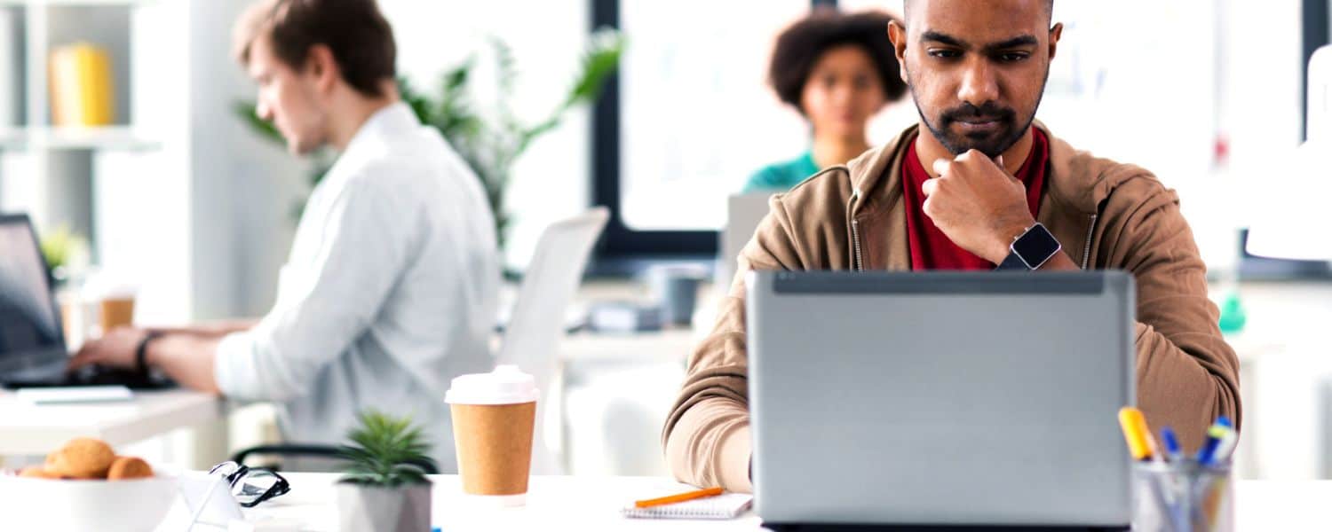 Male executive sits in modern office environment with laptop and takeaway coffee studying his screen looking perplexed. Two colleagues sit on separate desks behind him working on their laptops. Representing blog titled: Why your business needs a modern EDI platform