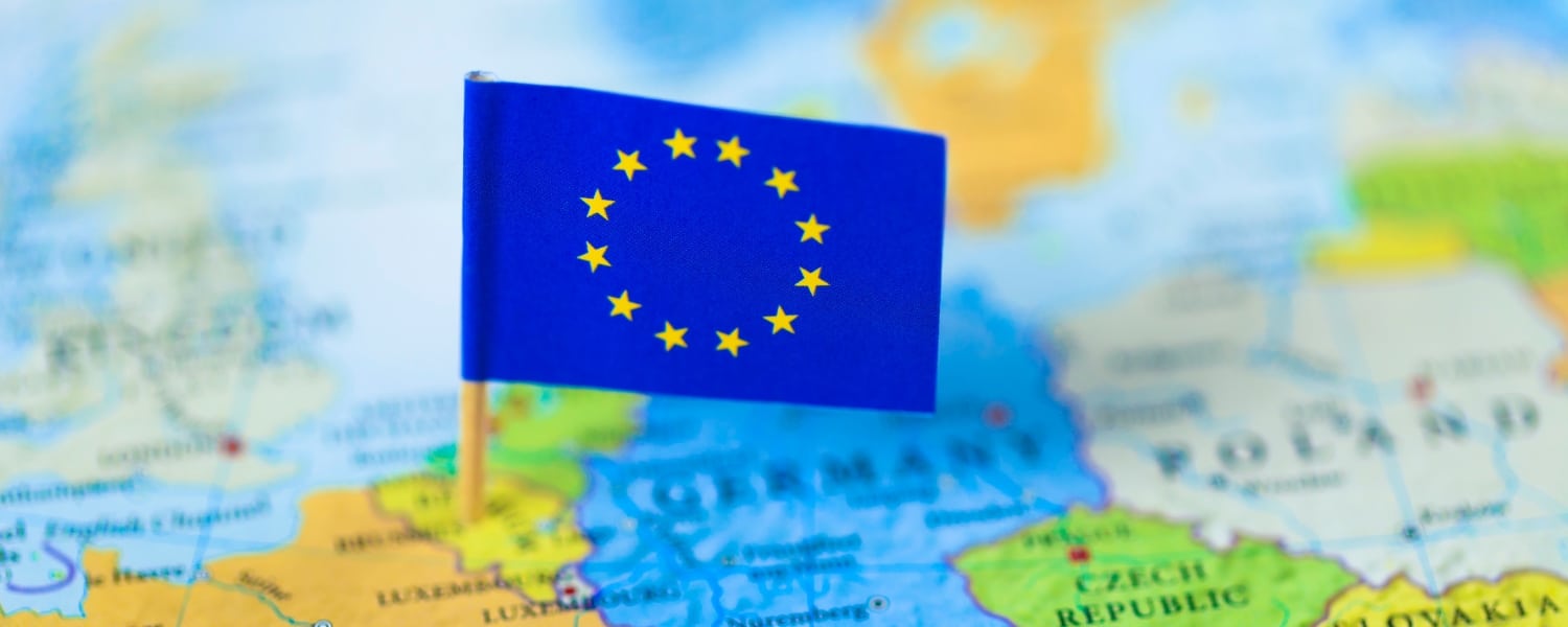 Out of focus map of the continent of Europe in full colour. Mini European flag of stars on cocktail stick in focus and pinned to the location of Brussels in Belgium. Representing the header image of a blog post by Transalis, titled: eInvoicing in Europe: Upcoming deadlines in 2026